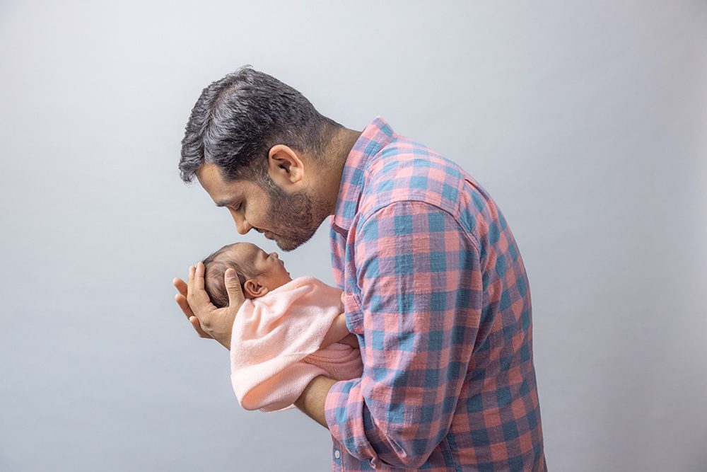 Father in plaid shirt tenderly holding wrapped newborn baby against gray background