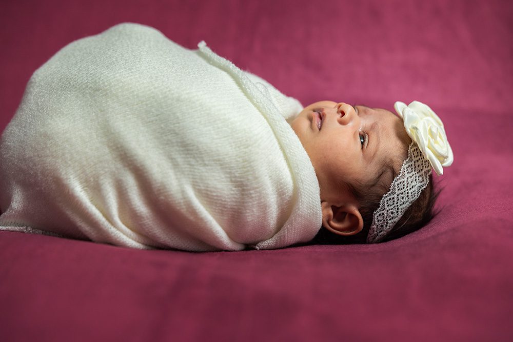 Alert newborn in cream wrap with white flower headband on pink textured background