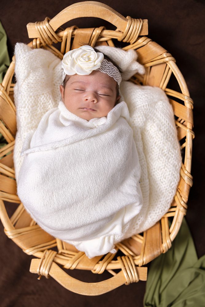 Sleeping newborn in white wrap with flower headband nestled in wicker basket