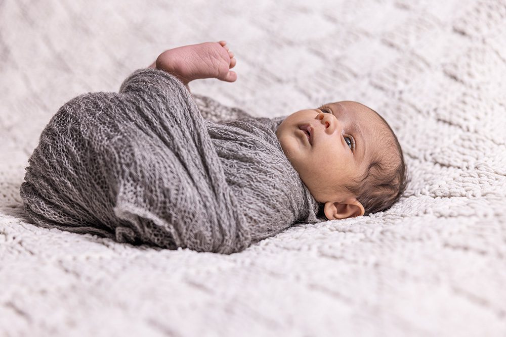 Alert newborn baby swaddled in charcoal gray wrap on white textured blanket