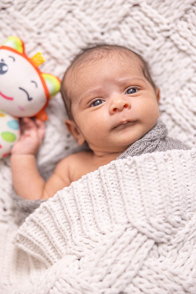 Awake newborn baby in gray wrap holding colorful rattle toy on white blanket