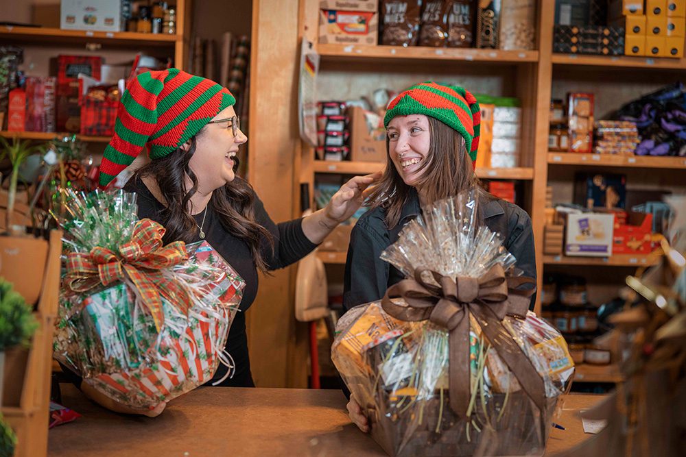 Two women in elf hats laughing while holding wrapped holiday gift baskets