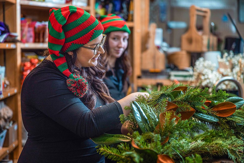 Two women in elf hats arranging evergreen garland with magnolia leaves in retail store