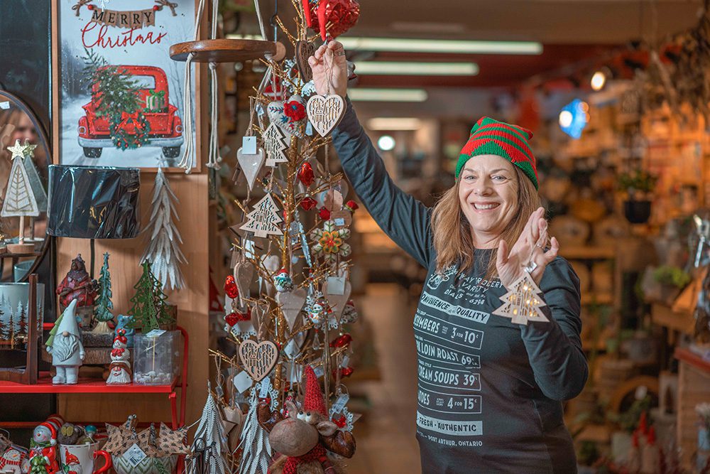 Woman in elf hat decorating rustic Christmas tree with ornaments in retail store