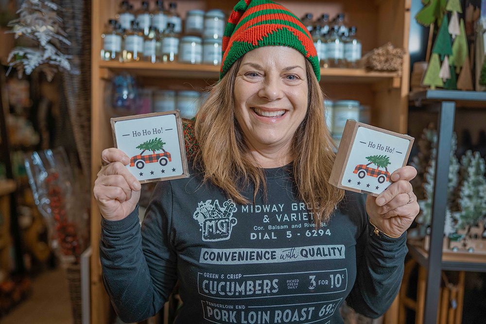 Smiling woman in red and green elf hat holding two "Ho Ho Ho" gift boxes at Georges Market