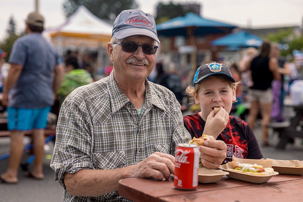 Senior man and young boy sitting at outdoor table with festival food and drinks at West Fort Street Fair
