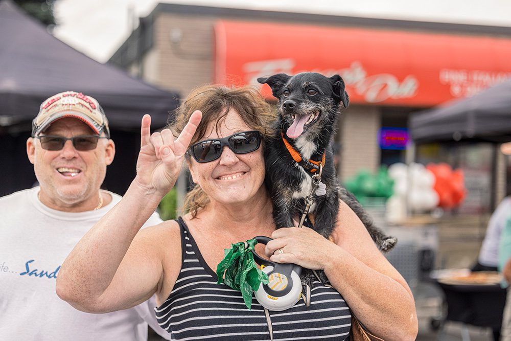 Woman holding small dog on shoulders making peace sign at community festival