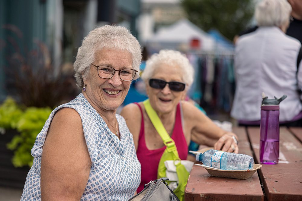 Two senior women sitting at outdoor table enjoying refreshments at community festival