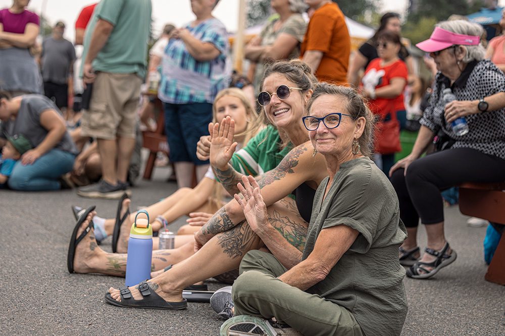 Two women sitting on street ground waving at camera during outdoor festival