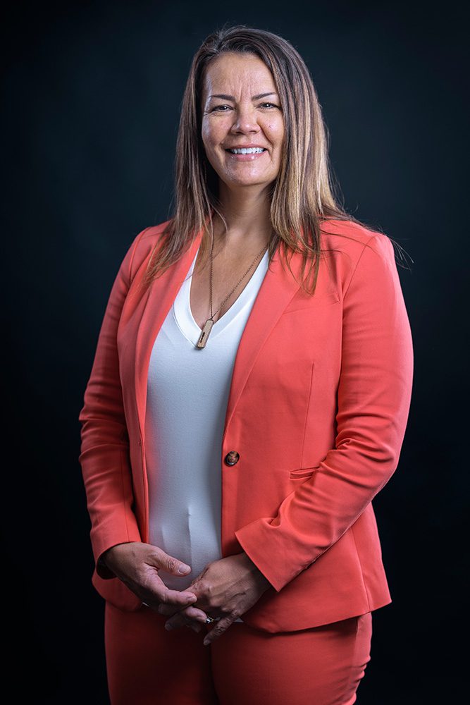 Professional woman in coral blazer with white top against dark background studio headshot