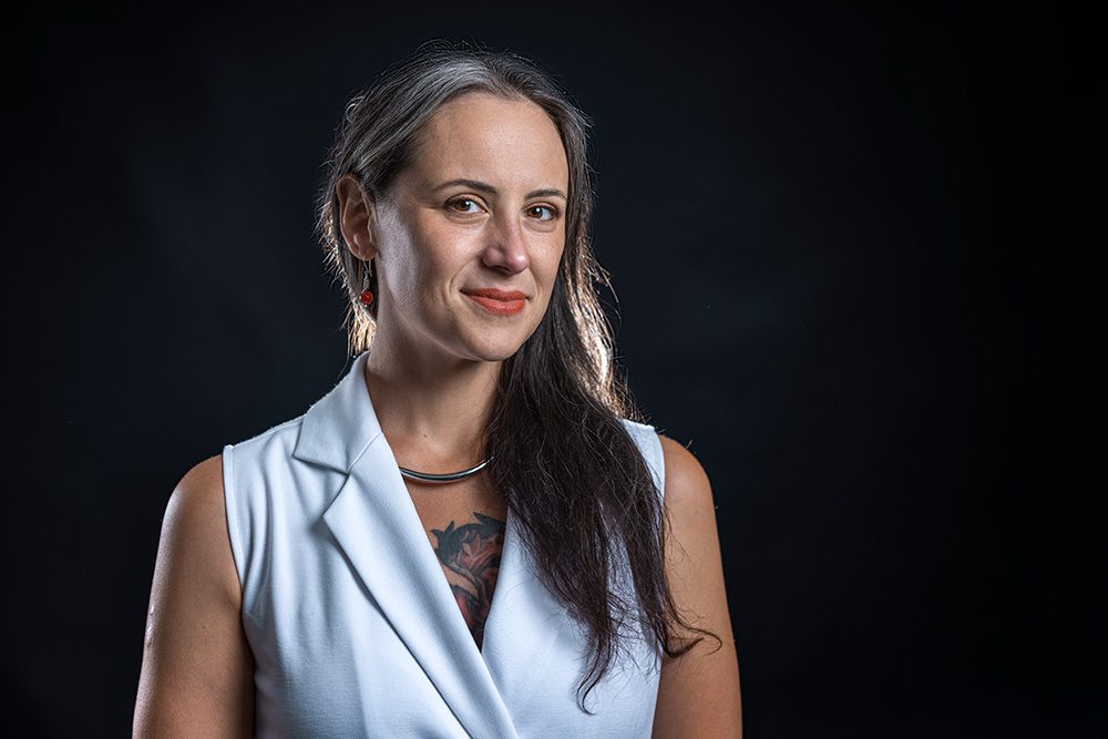 Professional woman with long dark hair in light blue sleeveless vest against black background